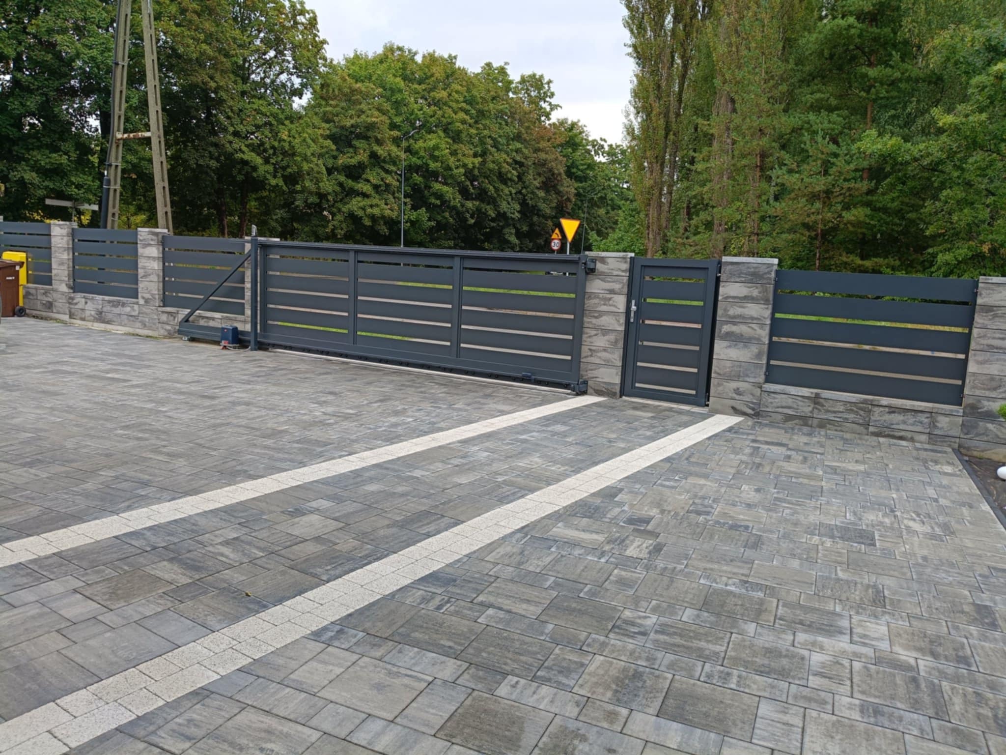 Nowoczesna, przesuwna brama w kolorze grafitowym z poziomymi panelami, osadzona na kamiennych słupkach. Ułożona kostka brukowa w odcieniach szarości. Tło: drzewa.