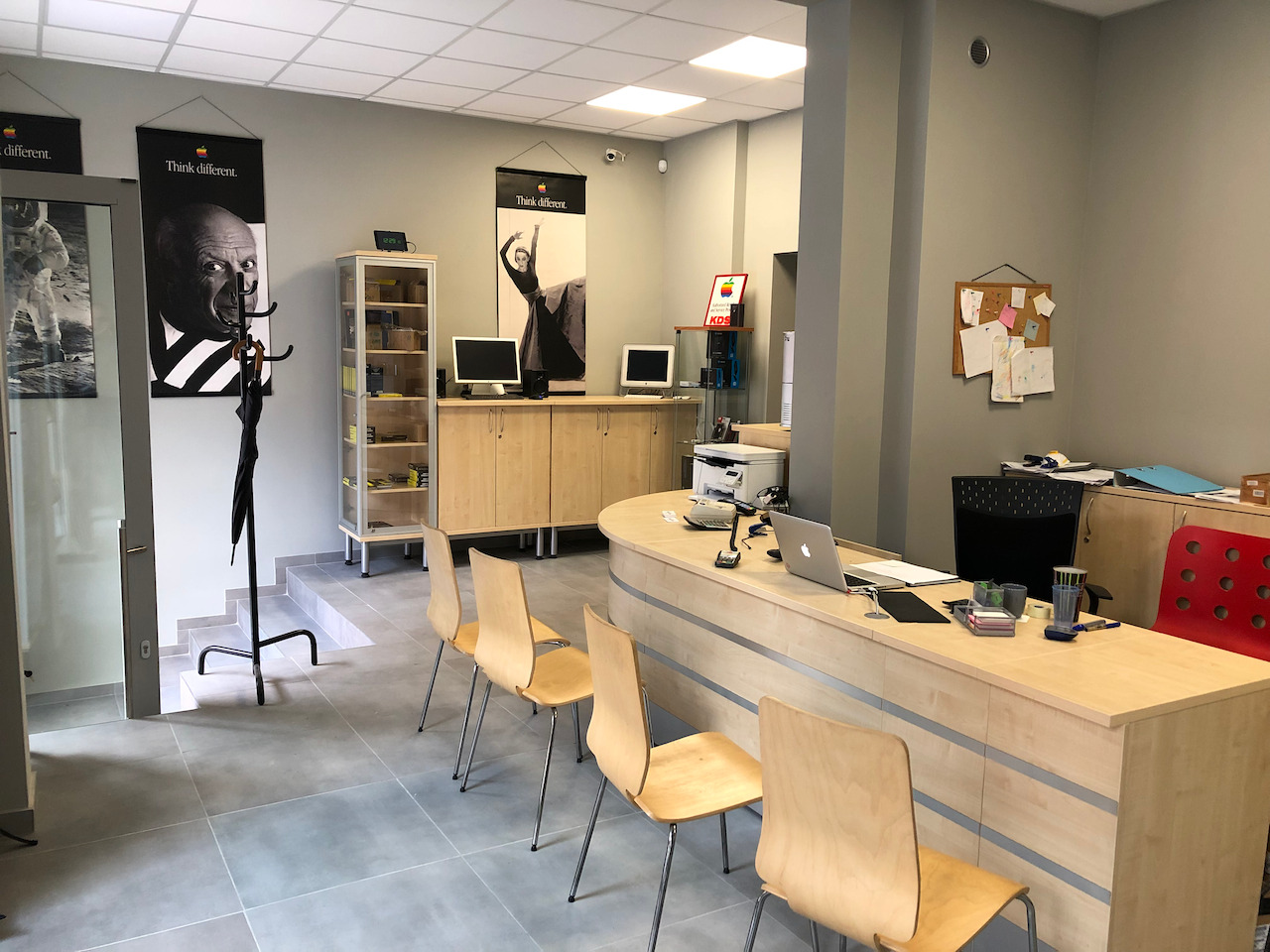 Wnętrze serwisu komputerowego z biurkiem obsługi, krzesłami, plakatami 'Think different' i sprzętem Apple. Widoczny komputer, drukarka i akcesoria.