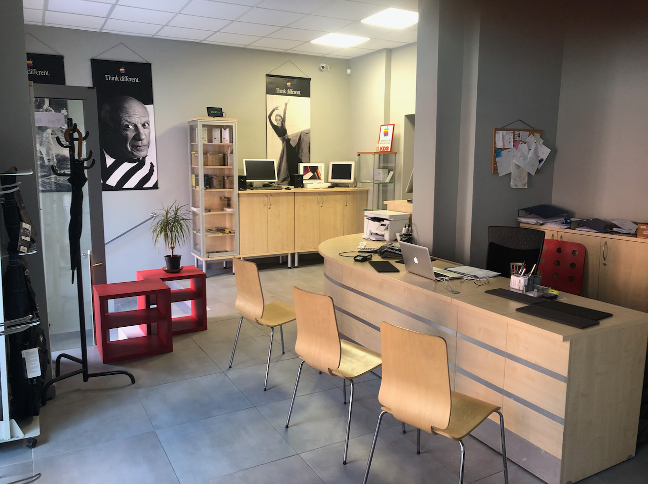 Wnętrze serwisu komputerowego z recepcją, sprzętem Apple, plakatami 'Think Different' i rośliną w doniczce. Widoczne monitory, laptop i akcesoria.