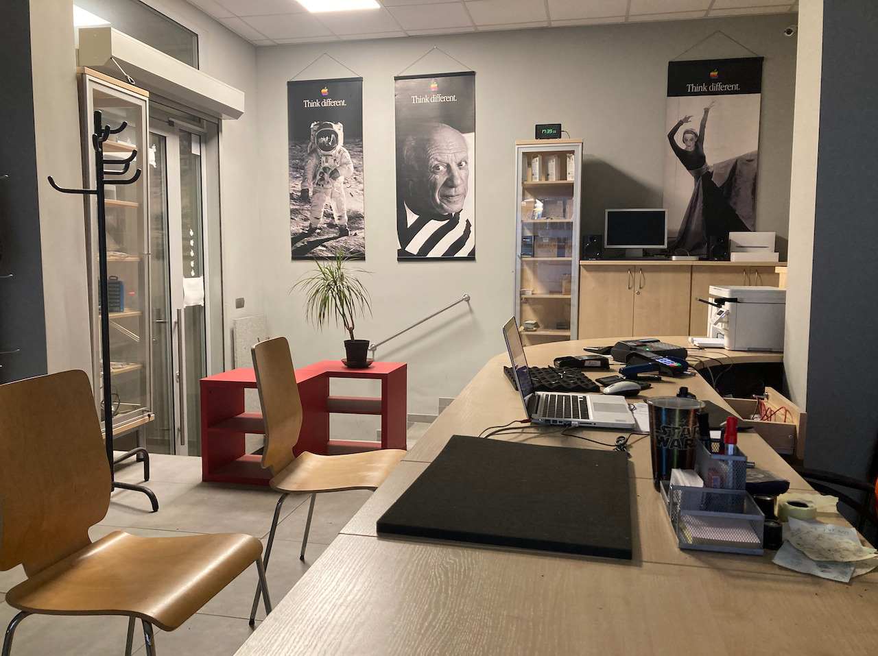 Biuro z plakatami 'Think different', biurkiem z laptopem, monitorem i akcesoriami. W tle regał z urządzeniami. Widok z perspektywy osoby stojącej przy biurku.