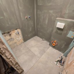 Aproxaa - Szpachlowana łazienka w trakcie remontu: ułożone szare płytki, widoczne instalacje, narzędzia. Ściany przygotowane pod malowanie, surowy stan wykończenia.