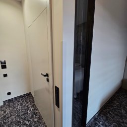 Aproxaa - Nowoczesne, białe drzwi z czarną klamką i panelem sterowania, obok szklanej ściany w pokoju z wzorzystą wykładziną. Minimalistyczny design.
