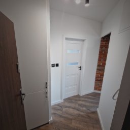 Aproxaa - Nowoczesny korytarz po remoncie: białe ściany, drzwi z przeszkleniami, drewnopodobna podłoga i dekoracyjna ściana z czerwonej cegły. Oświetlenie sufitowe.