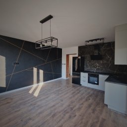 Aproxaa - Nowoczesna kuchnia z czarną ścianą akcentową i złotymi liniami, drewniana podłoga, stylowe oświetlenie. Widok na wykończone wnętrze po remoncie.