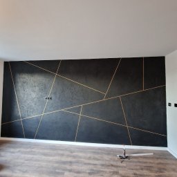 Aproxaa - Czarna ściana z geometrycznym wzorem w odcieniach złota, świeżo po malowaniu. Na podłodze widoczne narzędzia malarskie, podkreślające etap wykończenia wnętrza.