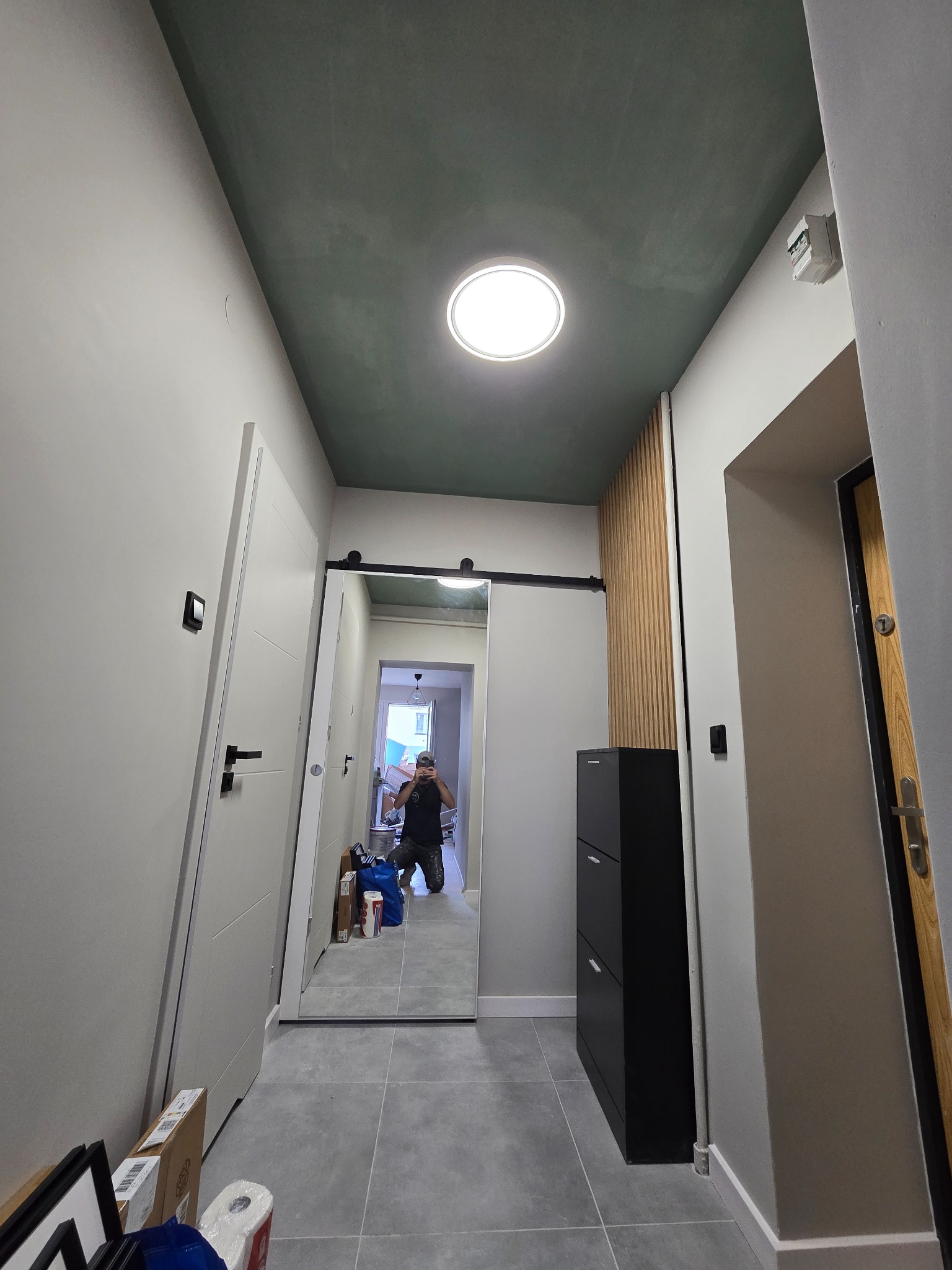 Nowoczesny przedpokój po remoncie: białe ściany, szary gres, ciemnozielony sufit z okrągłym oświetleniem, przesuwne drzwi i drewniane lamele.