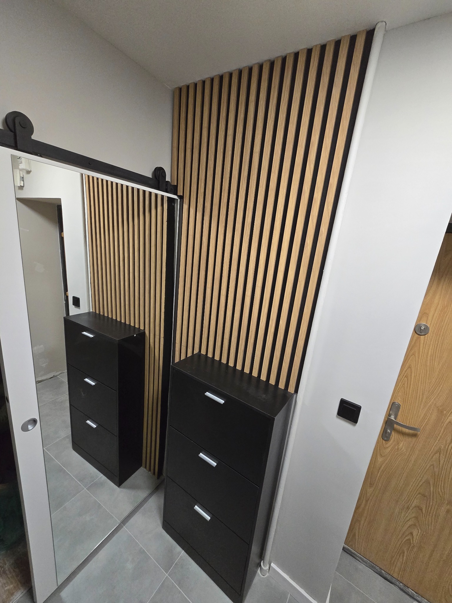 Nowoczesny przedpokój z drewnianą lamelową ścianą, czarnymi komodami i lustrem przesuwnym na szynie. Minimalistyczny design, szare płytki podłogowe.