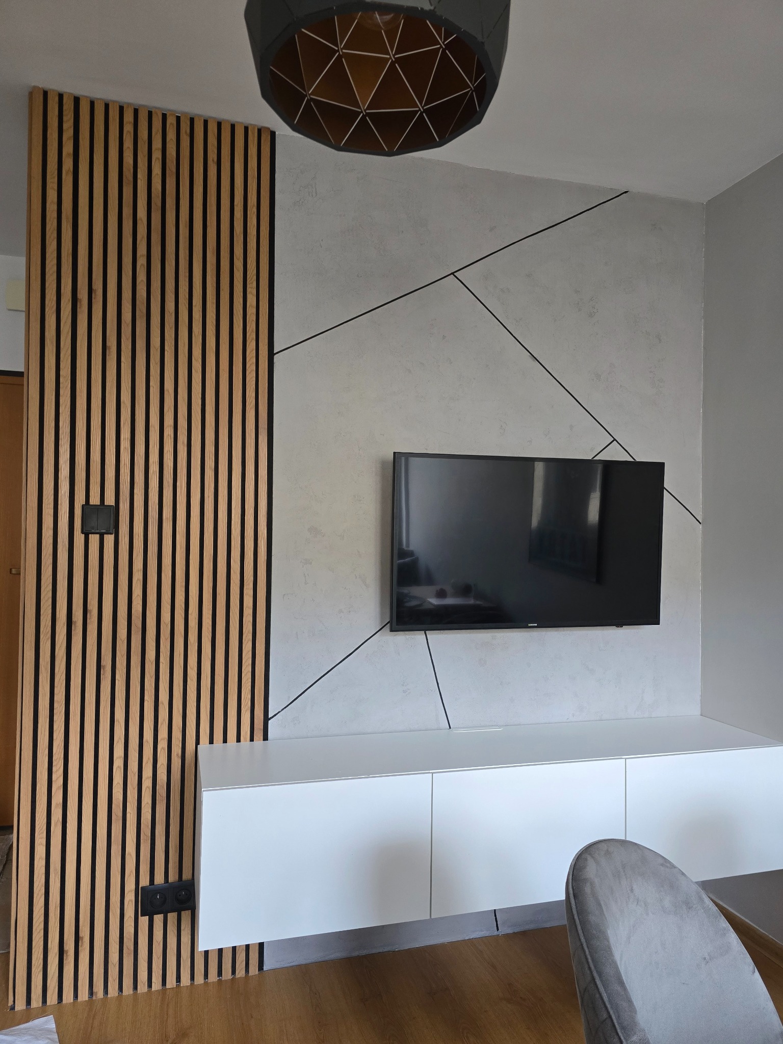 Nowoczesny salon z drewnianą ścianką działową, telewizorem na ścianie z geometrycznym wzorem i białą szafką RTV. Widoczna lampa i fragment krzesła.