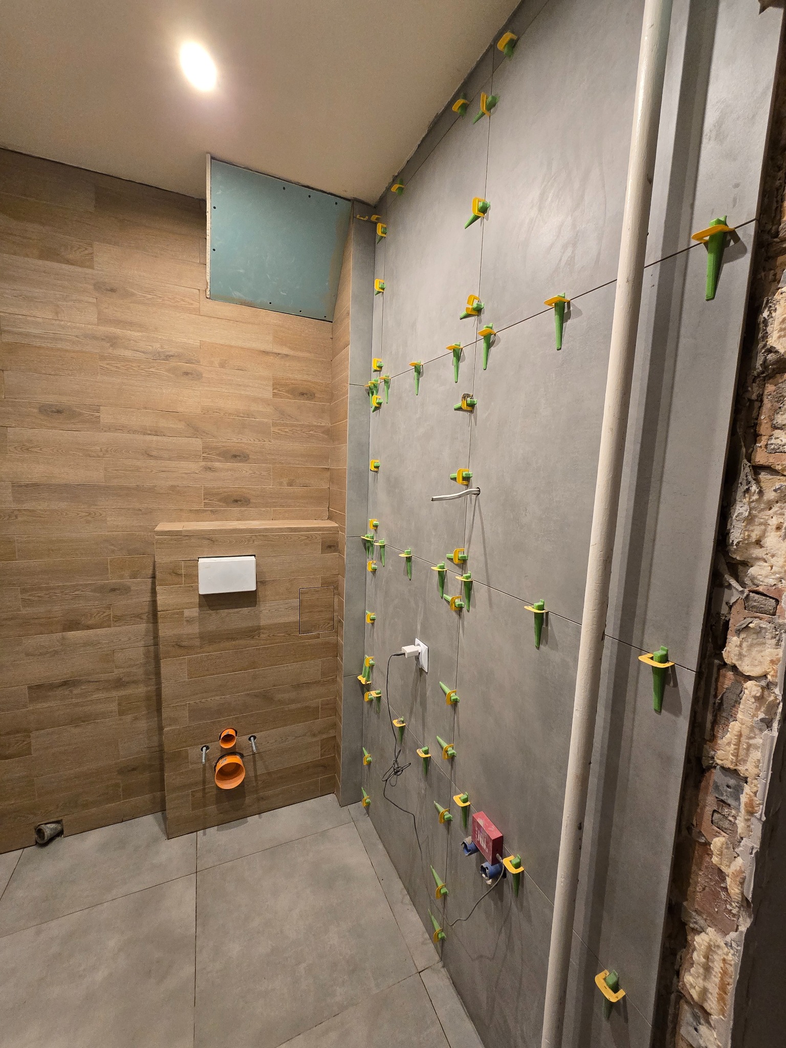 Łazienka w trakcie wykończenia: szare płytki z systemem poziomowania na ścianie, drewnopodobne płytki na zabudowie WC. Widoczne rury i fragment odsłoniętej cegły.