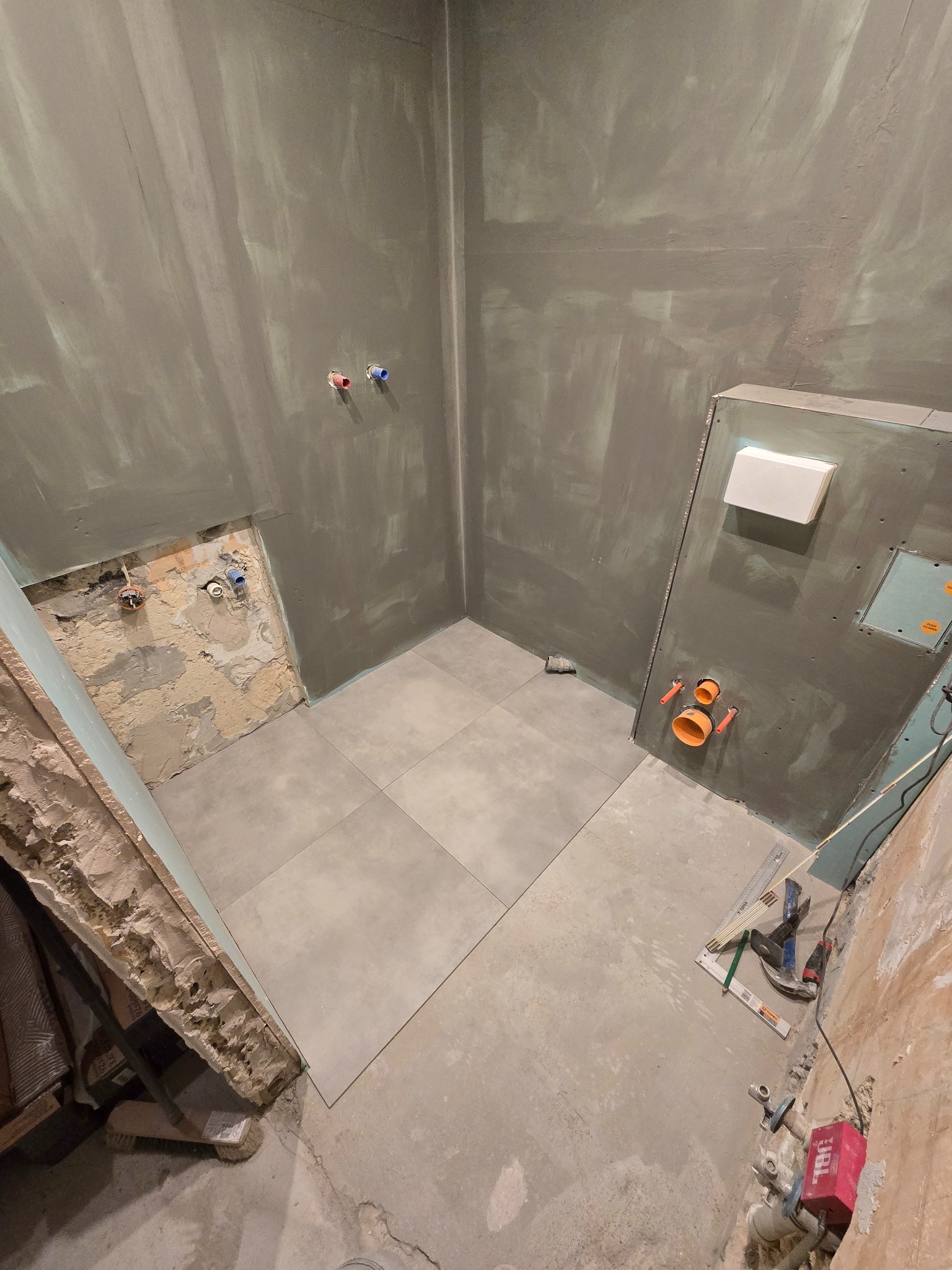 Szpachlowana łazienka w trakcie remontu: ułożone szare płytki, widoczne instalacje, narzędzia. Ściany przygotowane pod malowanie, surowy stan wykończenia.