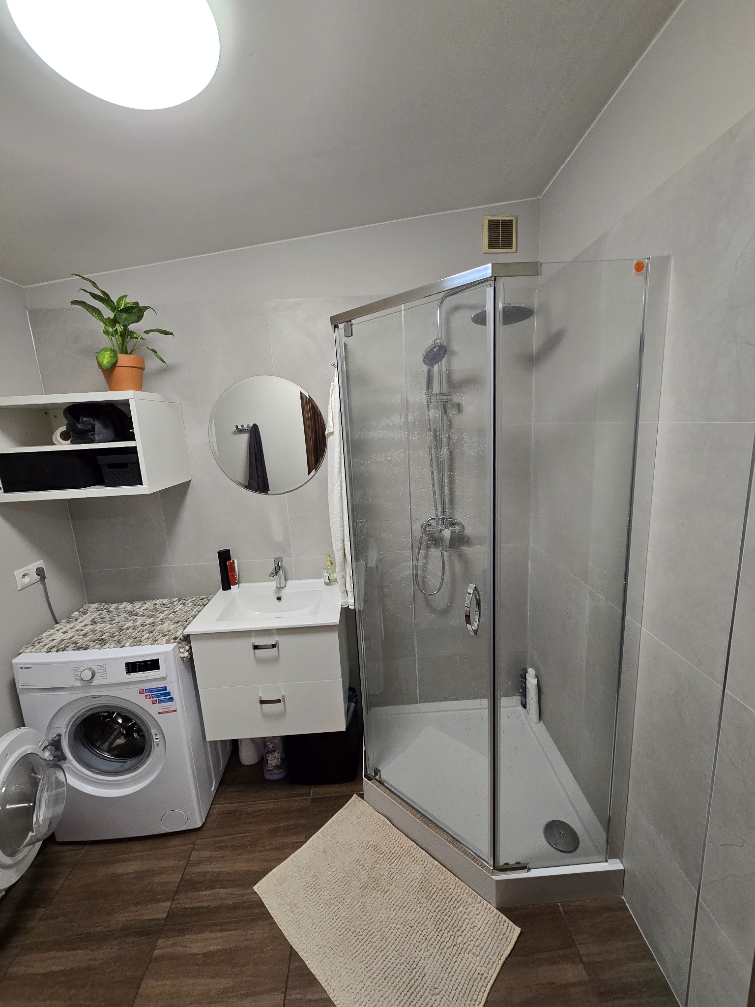 Nowoczesna łazienka w szarościach z kabiną prysznicową, pralką i rośliną doniczkową. Umywalka z szafką i okrągłe lustro dopełniają wnętrze. Widoczna kratka wentylacyjna.