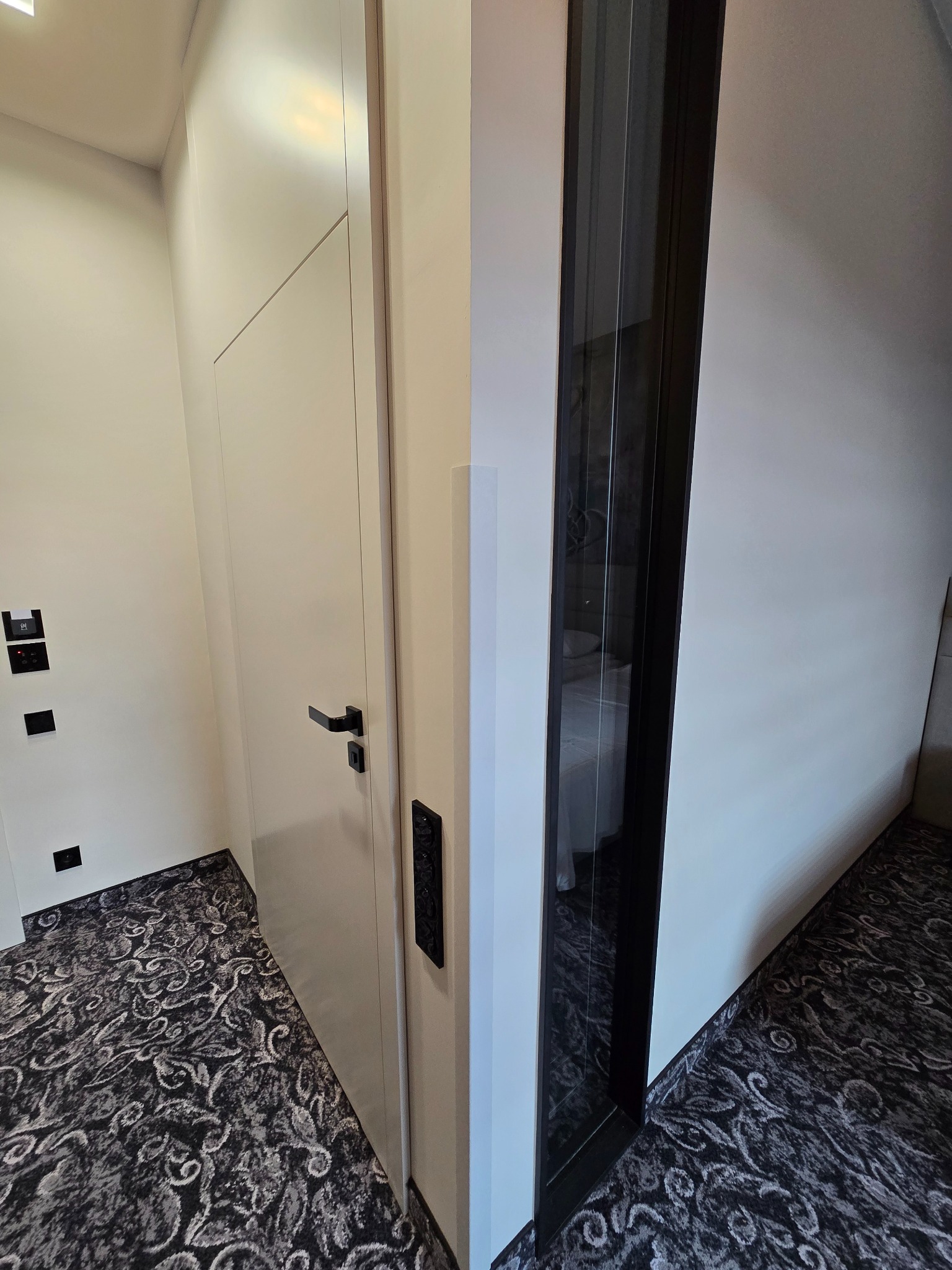 Nowoczesne, białe drzwi z czarną klamką i panelem sterowania, obok szklanej ściany w pokoju z wzorzystą wykładziną. Minimalistyczny design.