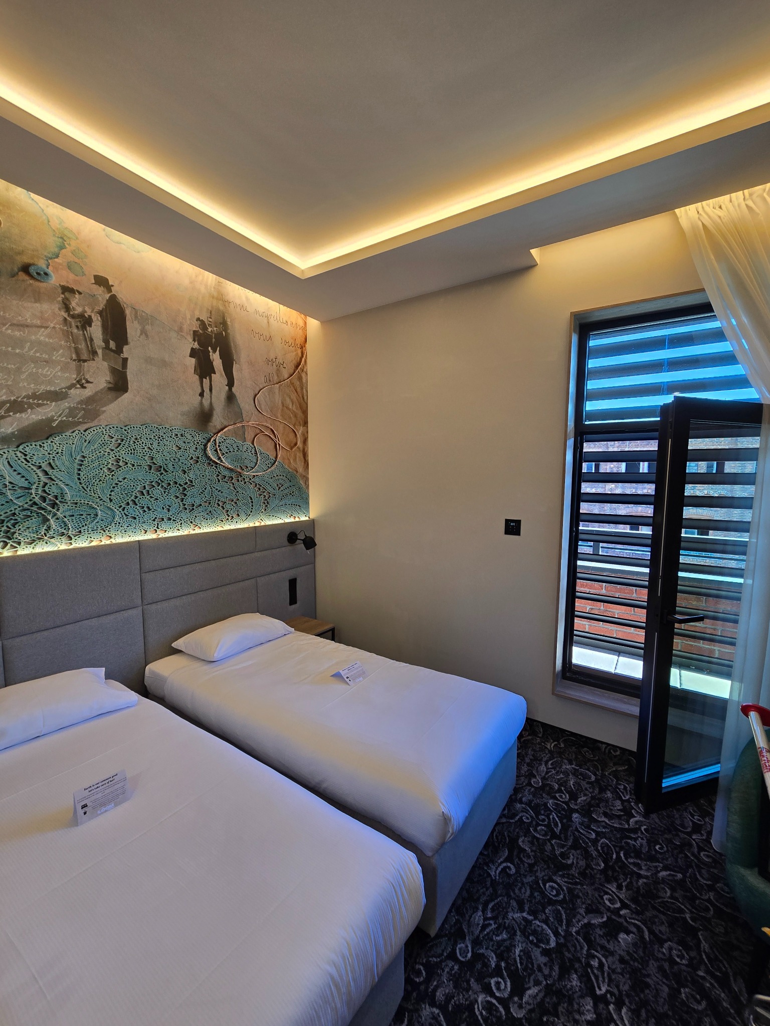 Pokój hotelowy z dwoma łóżkami pojedynczymi, dekoracyjną ścianą z motywem historycznym i otwartym oknem z widokiem na ceglany budynek.