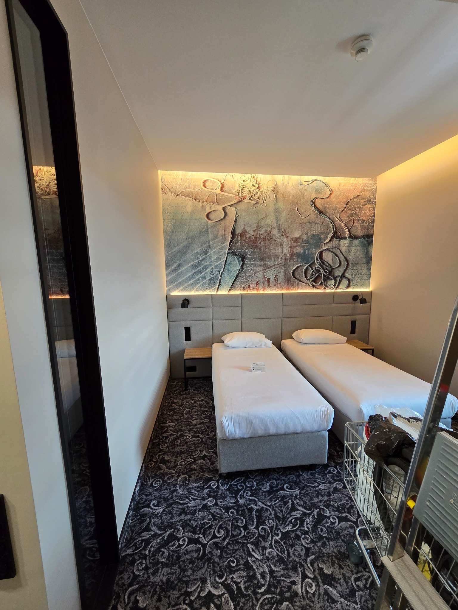 Pokój hotelowy z dwoma łóżkami pojedynczymi, wzorzystą tapetą i wózkiem serwisowym w rogu, ukazujący potencjalne prace wykończeniowe lub remontowe.