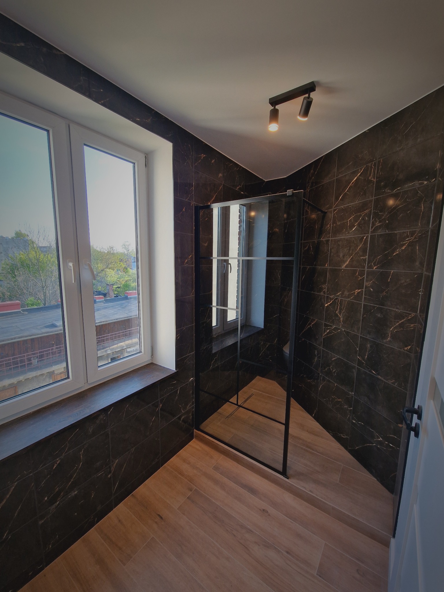 Nowoczesna łazienka z kabiną typu walk-in, wykończona płytkami imitującymi marmur w ciemnej tonacji i drewnopodobną podłogą. Widoczne okno z widokiem na zewnątrz.
