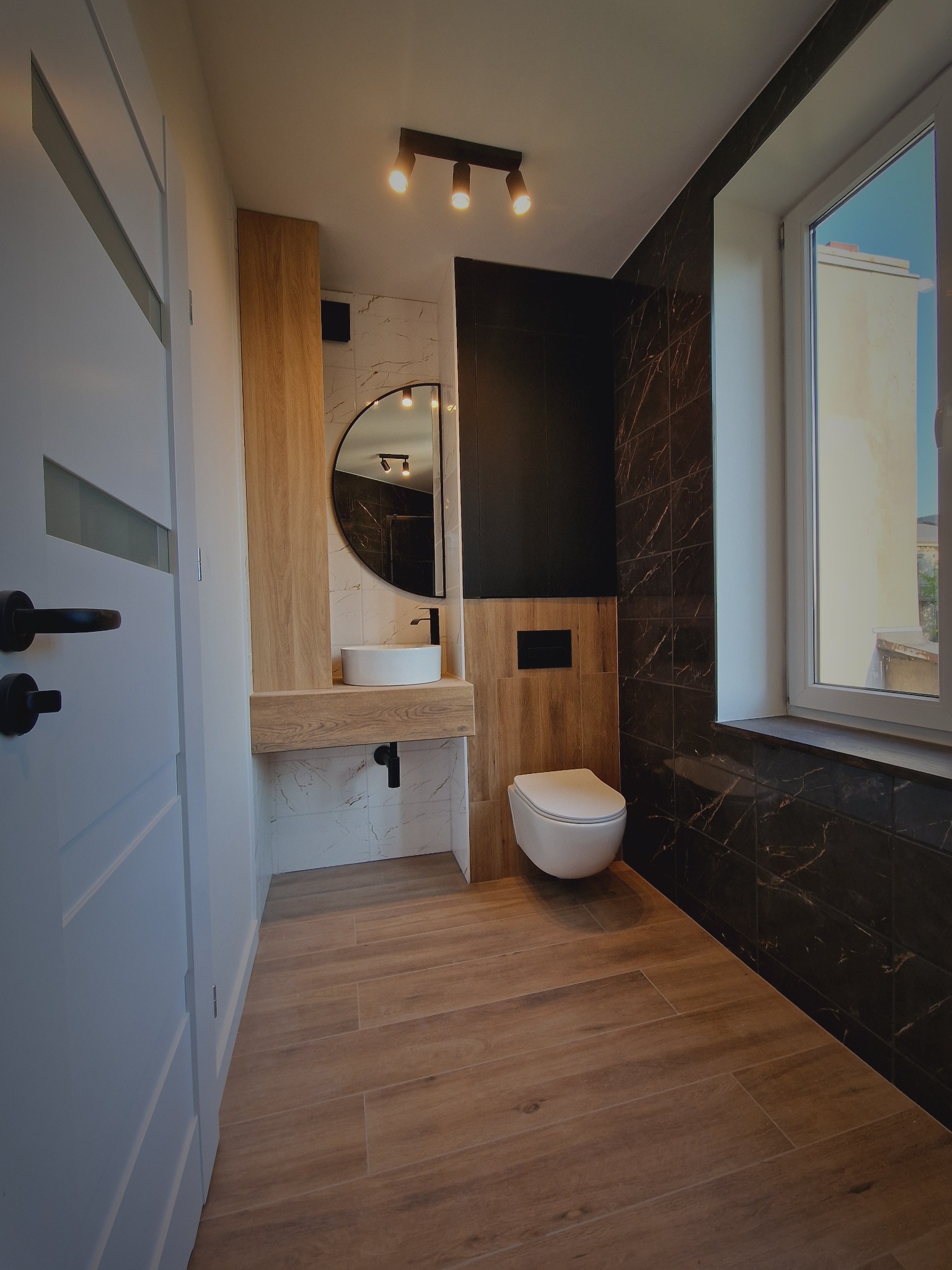 Nowoczesna łazienka z drewnianą podłogą i czarnymi płytkami imitującymi marmur. Umywalka na drewnianym blacie, okrągłe lustro i podwieszana toaleta.