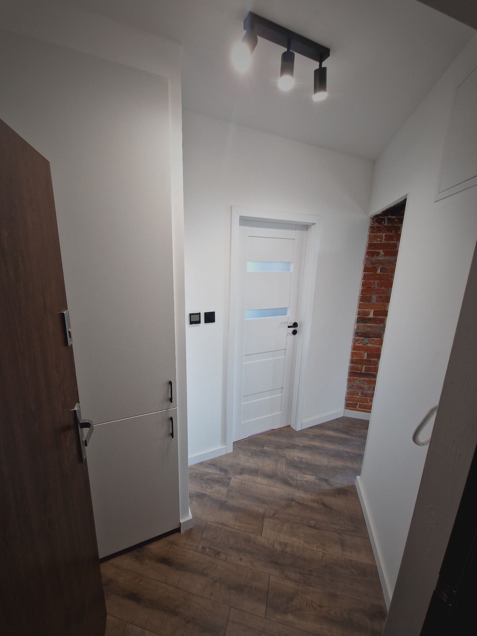 Nowoczesny korytarz po remoncie: białe ściany, drzwi z przeszkleniami, drewnopodobna podłoga i dekoracyjna ściana z czerwonej cegły. Oświetlenie sufitowe.