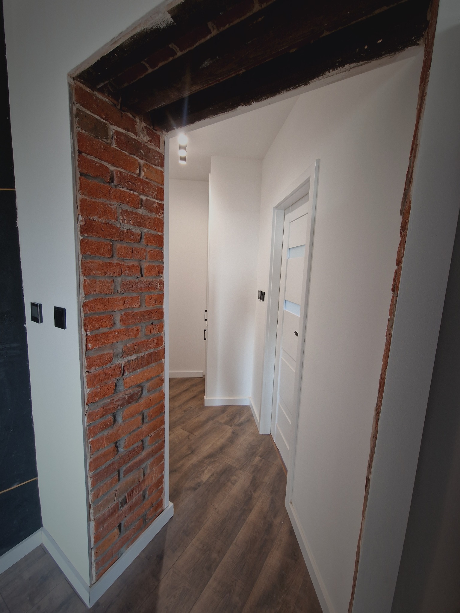 Wykończenie wnętrza: odnowione przejście z odsłoniętą czerwoną cegłą, kontrastujące z gładkimi, białymi ścianami i nowymi drzwiami z przeszkleniami. Nowoczesne oświetlenie.