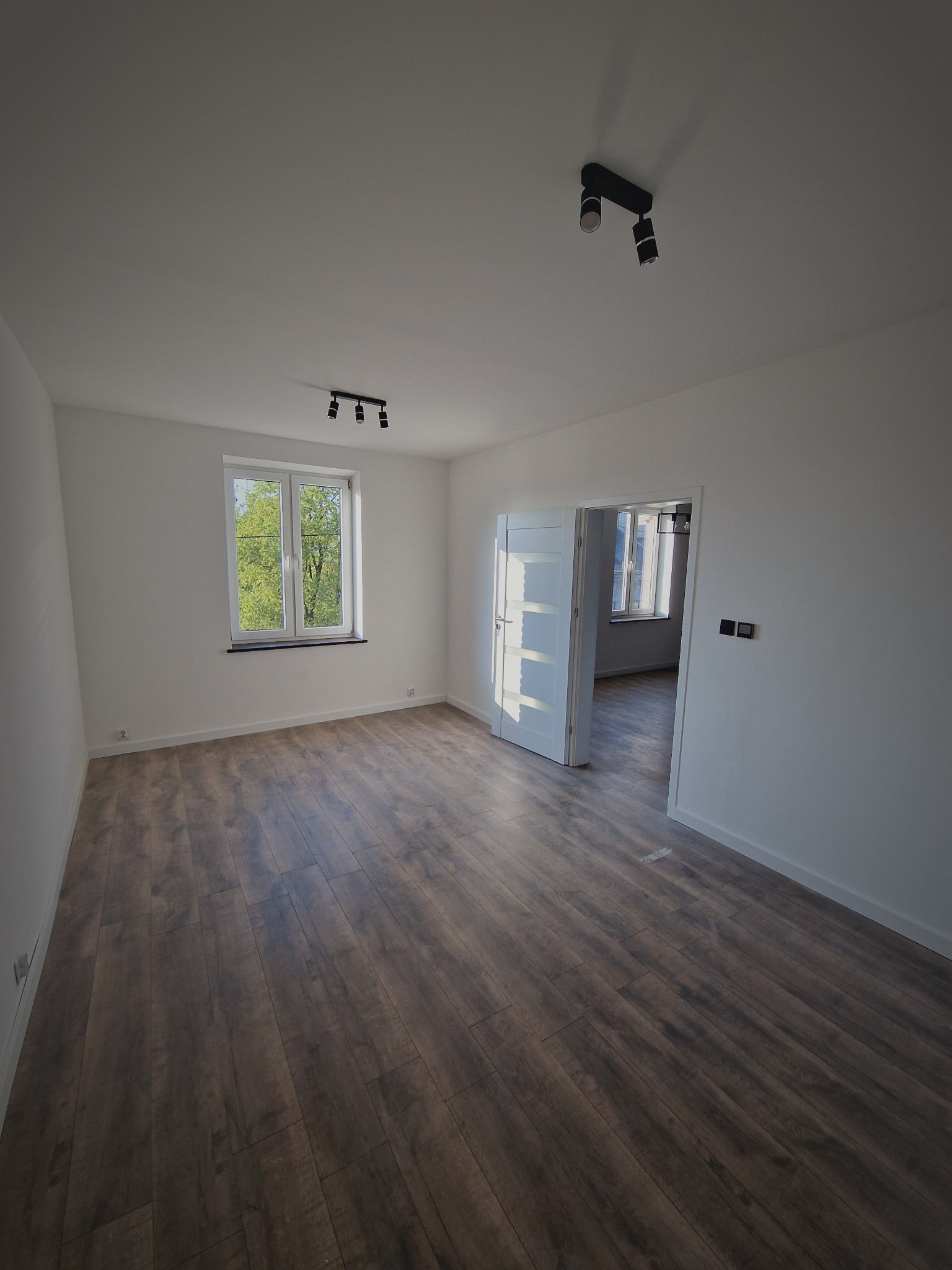 Jasne, przestronne wnętrze po remoncie: ciemne panele podłogowe, białe ściany, okno z widokiem na zieleń i otwarte, białe drzwi do kolejnego pomieszczenia. Minimalistyczne oświetlenie.