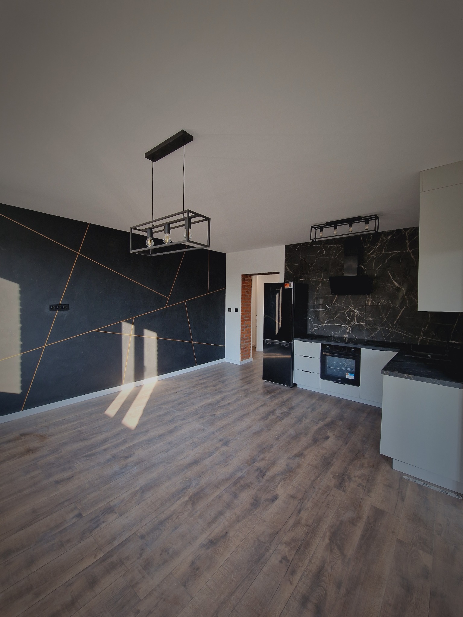 Nowoczesna kuchnia z czarną ścianą akcentową i złotymi liniami, drewniana podłoga, stylowe oświetlenie. Widok na wykończone wnętrze po remoncie.