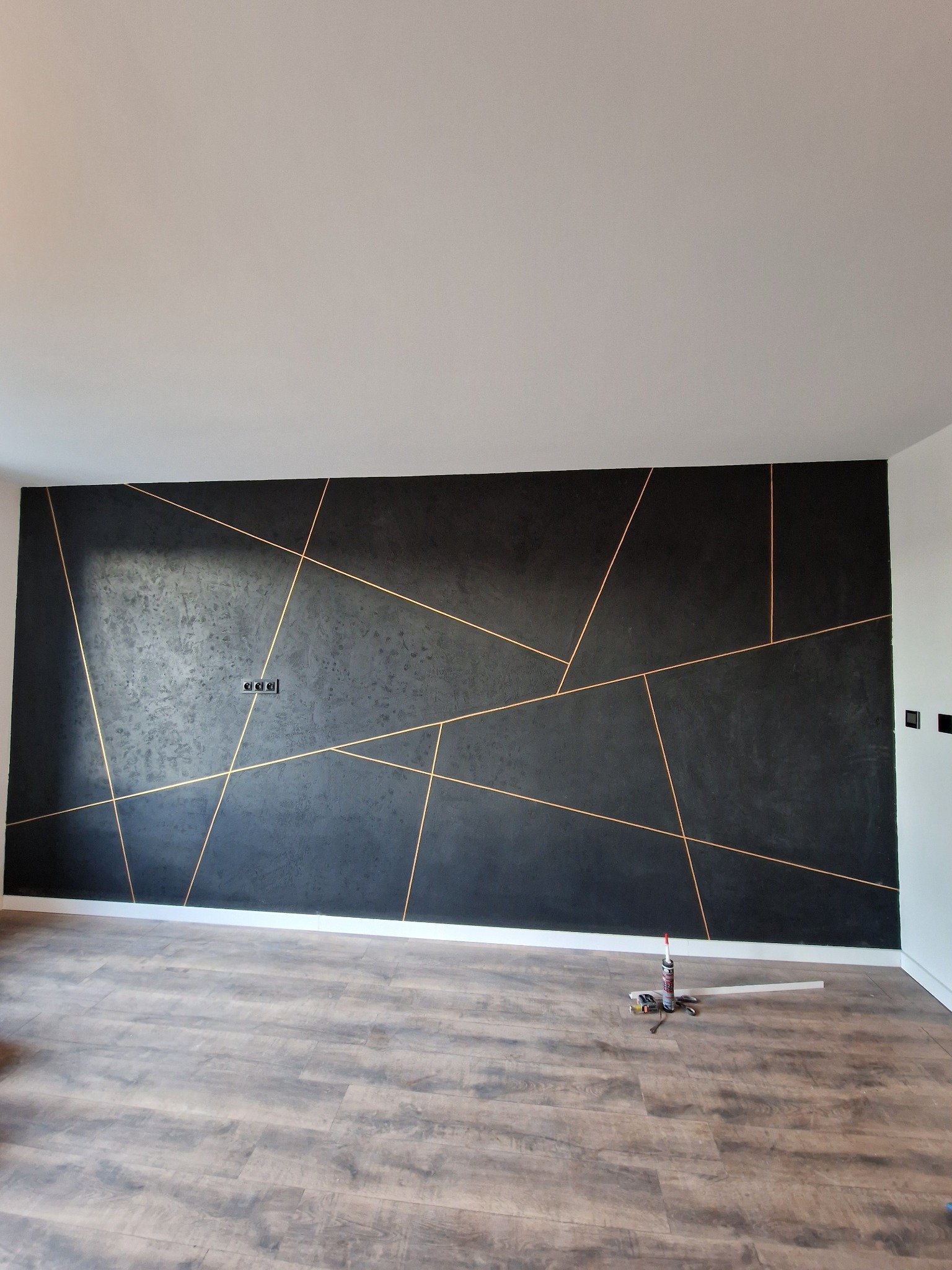 Czarna ściana z geometrycznym wzorem w odcieniach złota, świeżo po malowaniu. Na podłodze widoczne narzędzia malarskie, podkreślające etap wykończenia wnętrza.