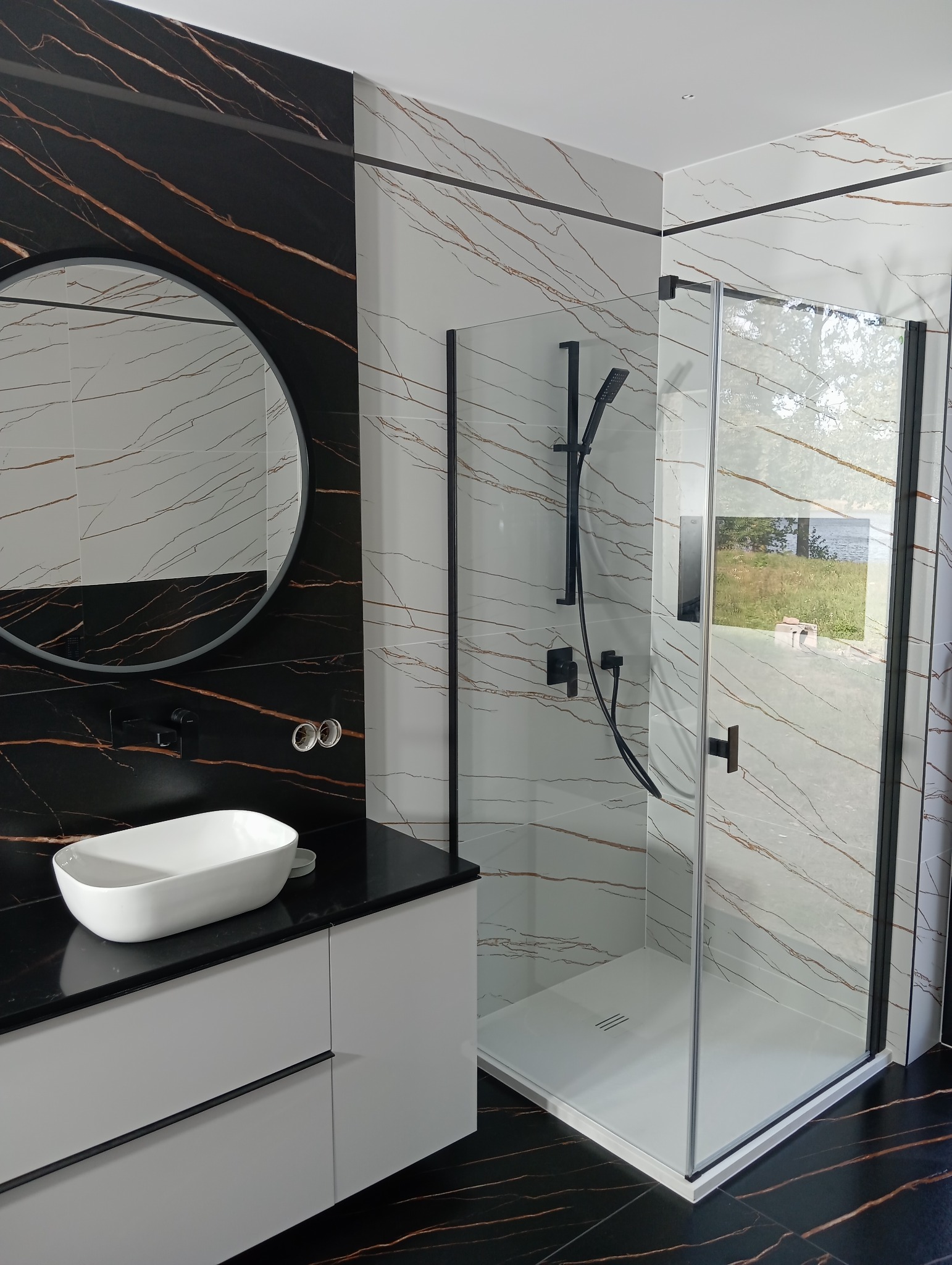 Nowoczesna łazienka z kabiną prysznicową typu walk-in, umywalką nablatową i okrągłym lustrem. Czarno-białe płytki imitujące marmur z brązowymi żyłami.