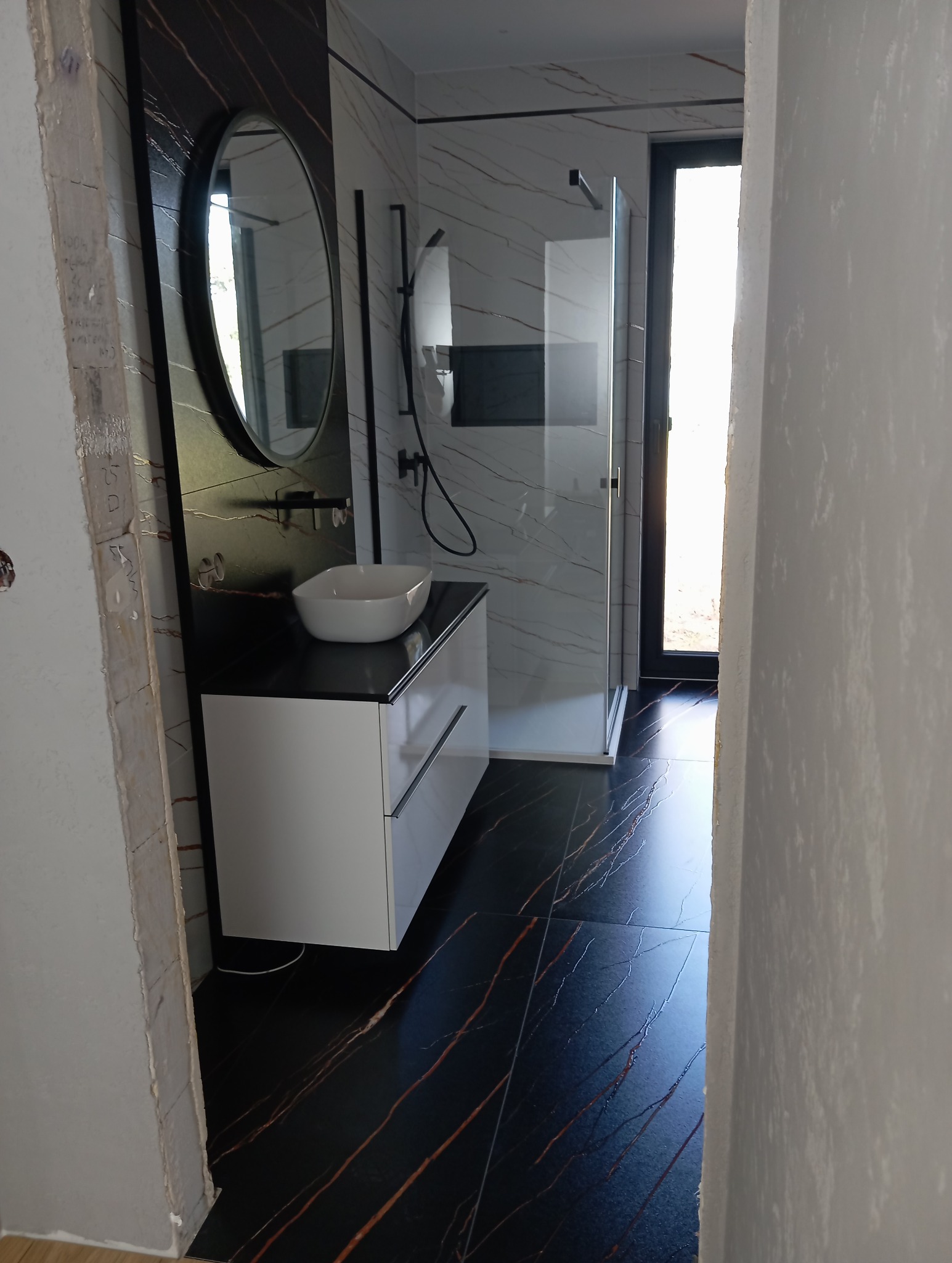 Nowoczesna łazienka z czarną podłogą w marmurkowy wzór, białą szafką pod umywalką i kabiną prysznicową. Widoczny fragment ściany w stanie surowym.