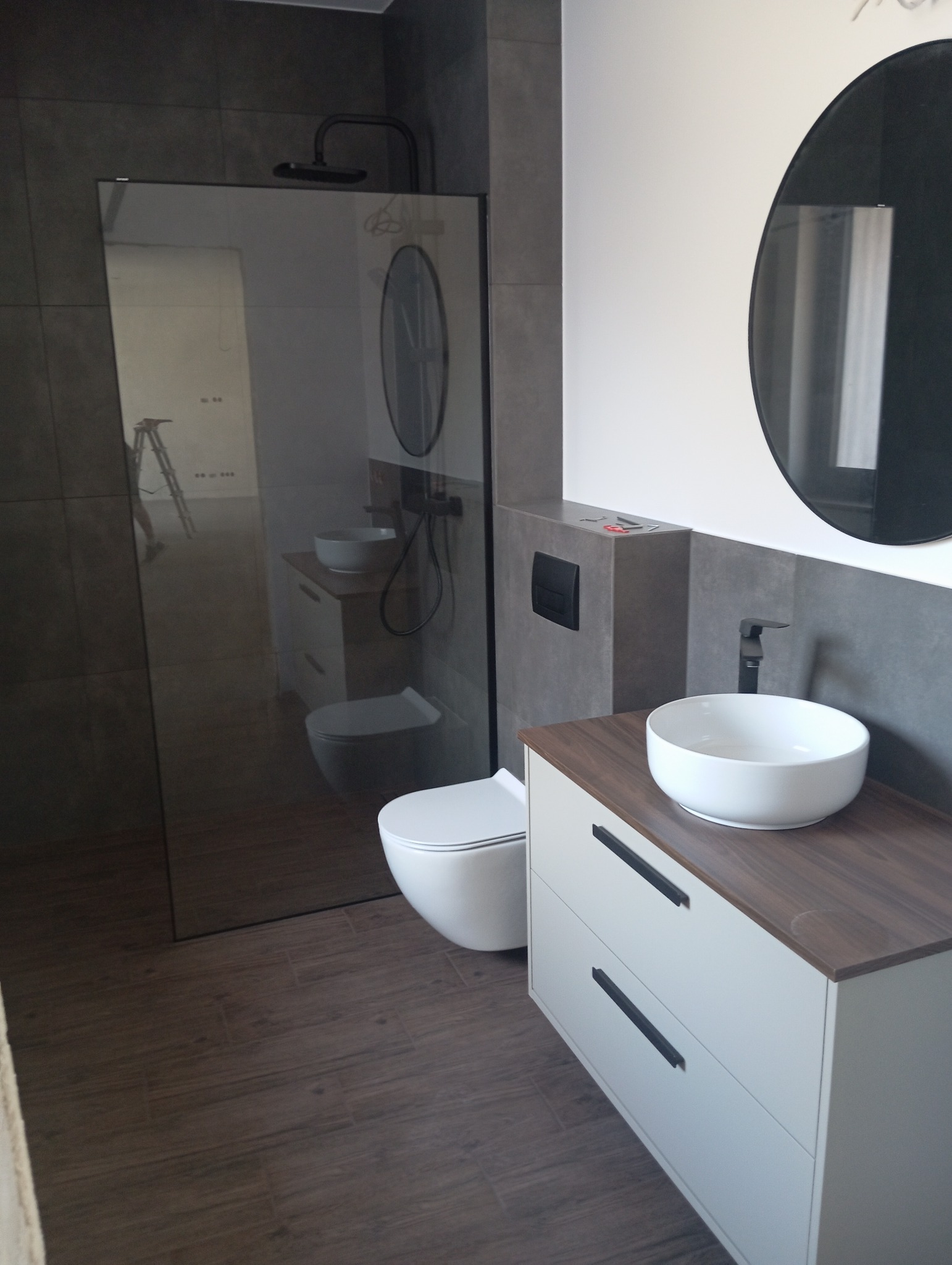 Nowoczesna łazienka z kabiną typu walk-in, umywalką nablatową na szafce z szufladami i okrągłym lustrem. Ciemne płytki i drewnopodobna podłoga.