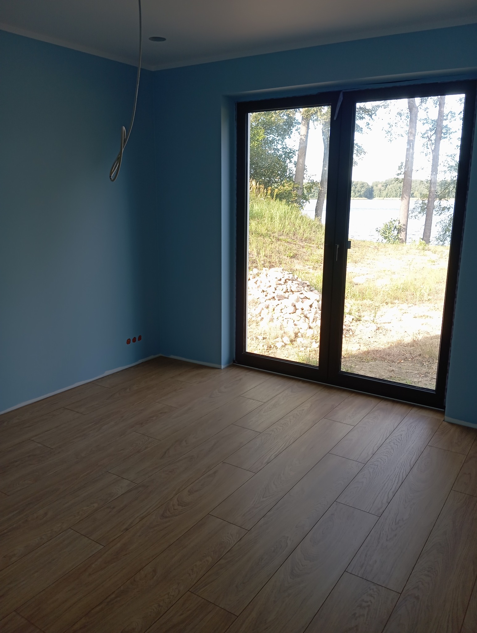 Błękitny pokój z drewnianą podłogą i dużym oknem z widokiem na jezioro. Widoczne gniazdka elektryczne i kabel zwisający z sufitu.