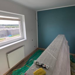 FINITO PAWEŁ GOWIN - Przygotowania do malowania pokoju: zabezpieczone meble folią, podłoga przykryta, widoczna gąbka i tuba z uszczelniaczem. Ściana pomalowana na niebiesko.