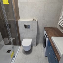 FINITO PAWEŁ GOWIN - Wykończona łazienka z kabiną prysznicową, podwieszaną toaletą i szafką z umywalką. Szare płytki na ścianach i podłodze. Widoczna pralka pod blatem.