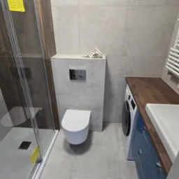 Wykończona łazienka z kabiną prysznicową, podwieszaną toaletą i szafką z umywalką. Szare płytki na ścianach i podłodze. Widoczna pralka pod blatem.