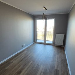 FINITO PAWEŁ GOWIN - Jasne, przestronne wnętrze z drewnianą podłogą, szarymi ścianami i balkonem. Nowoczesna lampa wisząca dodaje elegancji. Widok na zewnątrz przez przeszklone drzwi balkonowe.