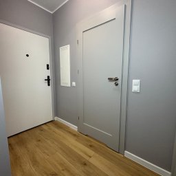 FINITO PAWEŁ GOWIN - Szare ściany, drewnopodobne panele i dwie białe, nowoczesne drzwi wewnętrzne z srebrnymi klamkami w przedpokoju po remoncie.