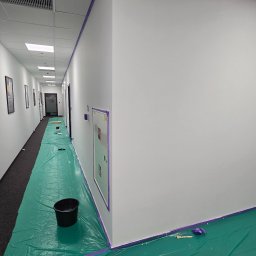 FINITO PAWEŁ GOWIN - Korytarz biurowy w trakcie malowania: zabezpieczona podłoga folią, świeżo pomalowane ściany na biało, taśma malarska wzdłuż krawędzi. Widoczny kubeł z farbą i drabina.