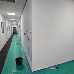 Korytarz biurowy w trakcie malowania: zabezpieczona podłoga folią, świeżo pomalowane ściany na biało, taśma malarska wzdłuż krawędzi. Widoczny kubeł z farbą i drabina.