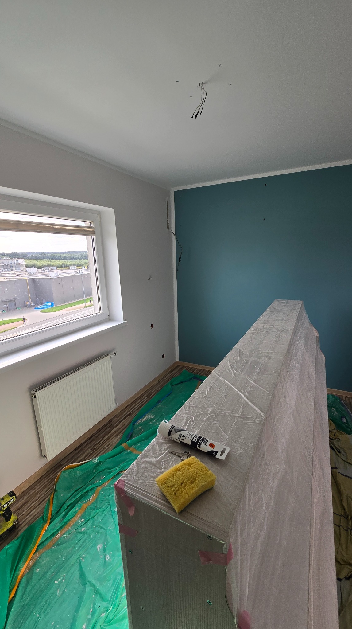 Przygotowania do malowania pokoju: zabezpieczone meble folią, podłoga przykryta, widoczna gąbka i tuba z uszczelniaczem. Ściana pomalowana na niebiesko.