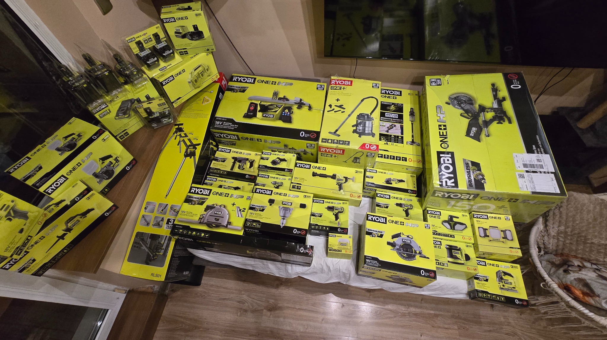 Zestaw narzędzi Ryobi One+ HP w oryginalnych opakowaniach, ułożonych na podłodze. Widoczne wiertarki, piły, odkurzacz i inne elektronarzędzia.