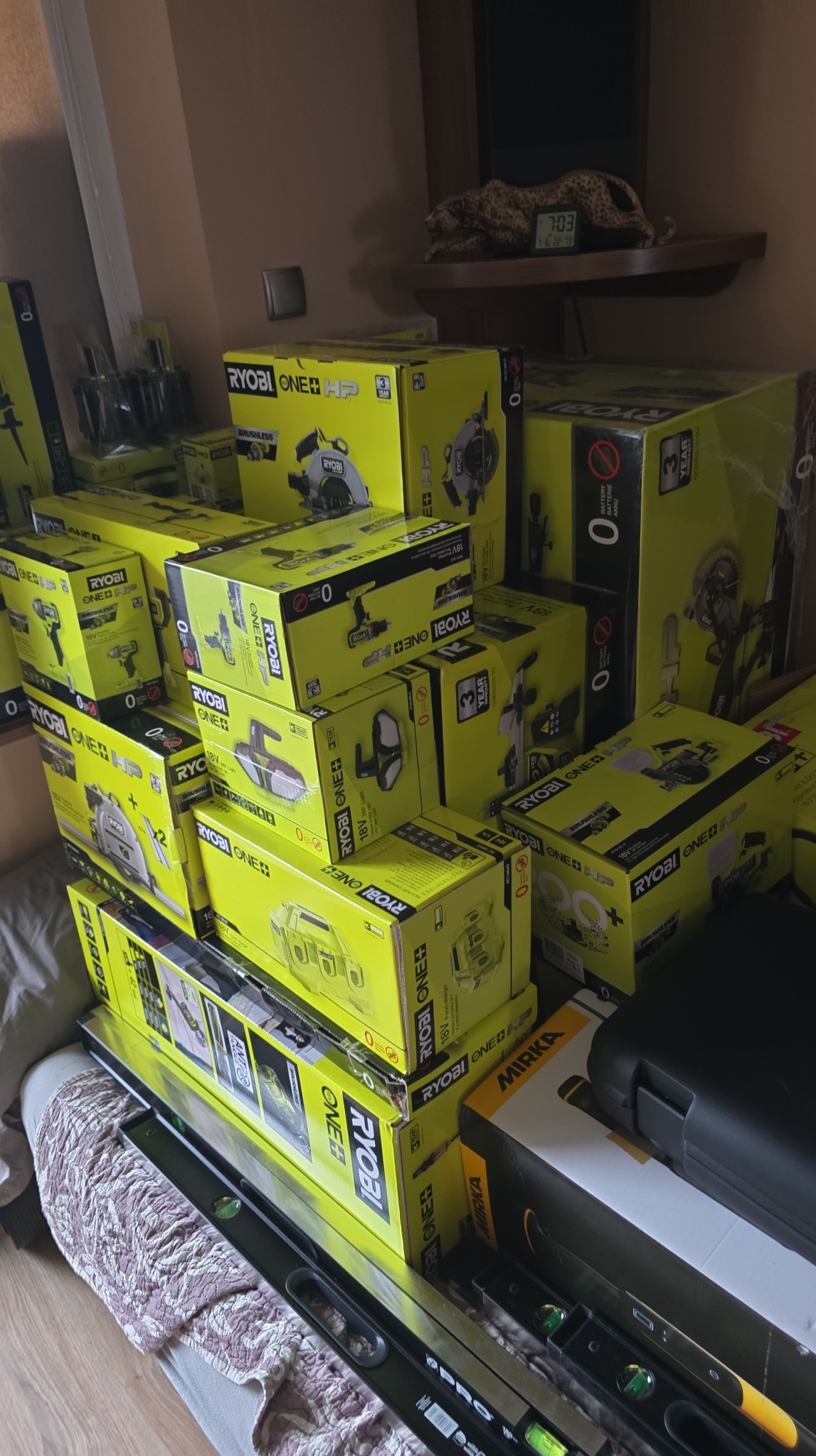 Stos żółto-czarnych pudełek Ryobi One+ z narzędziami, poziomice PRO i Mirka, na tle ściany i półki z figurką geparda i zegarem.