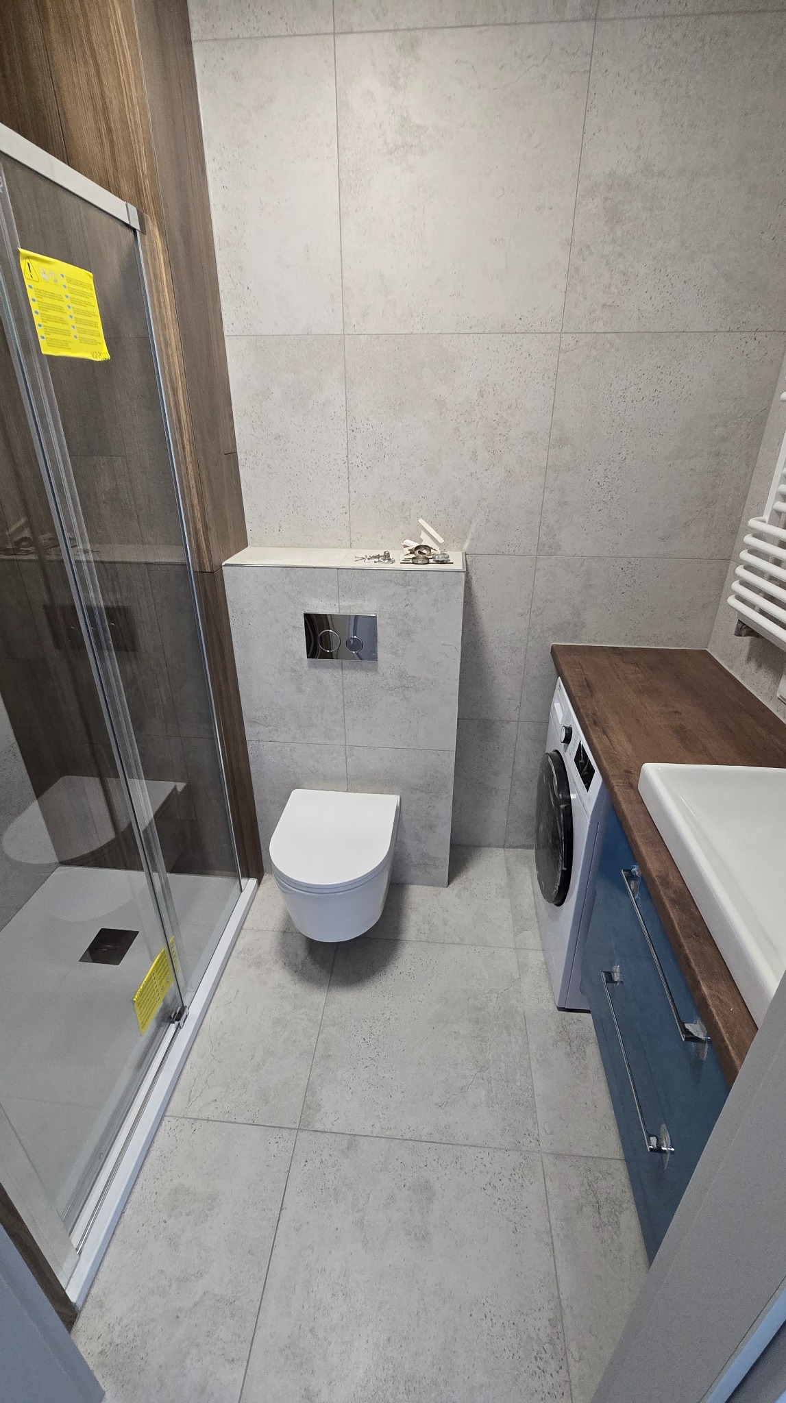 Wykończona łazienka z kabiną prysznicową, podwieszaną toaletą i szafką z umywalką. Szare płytki na ścianach i podłodze. Widoczna pralka pod blatem.