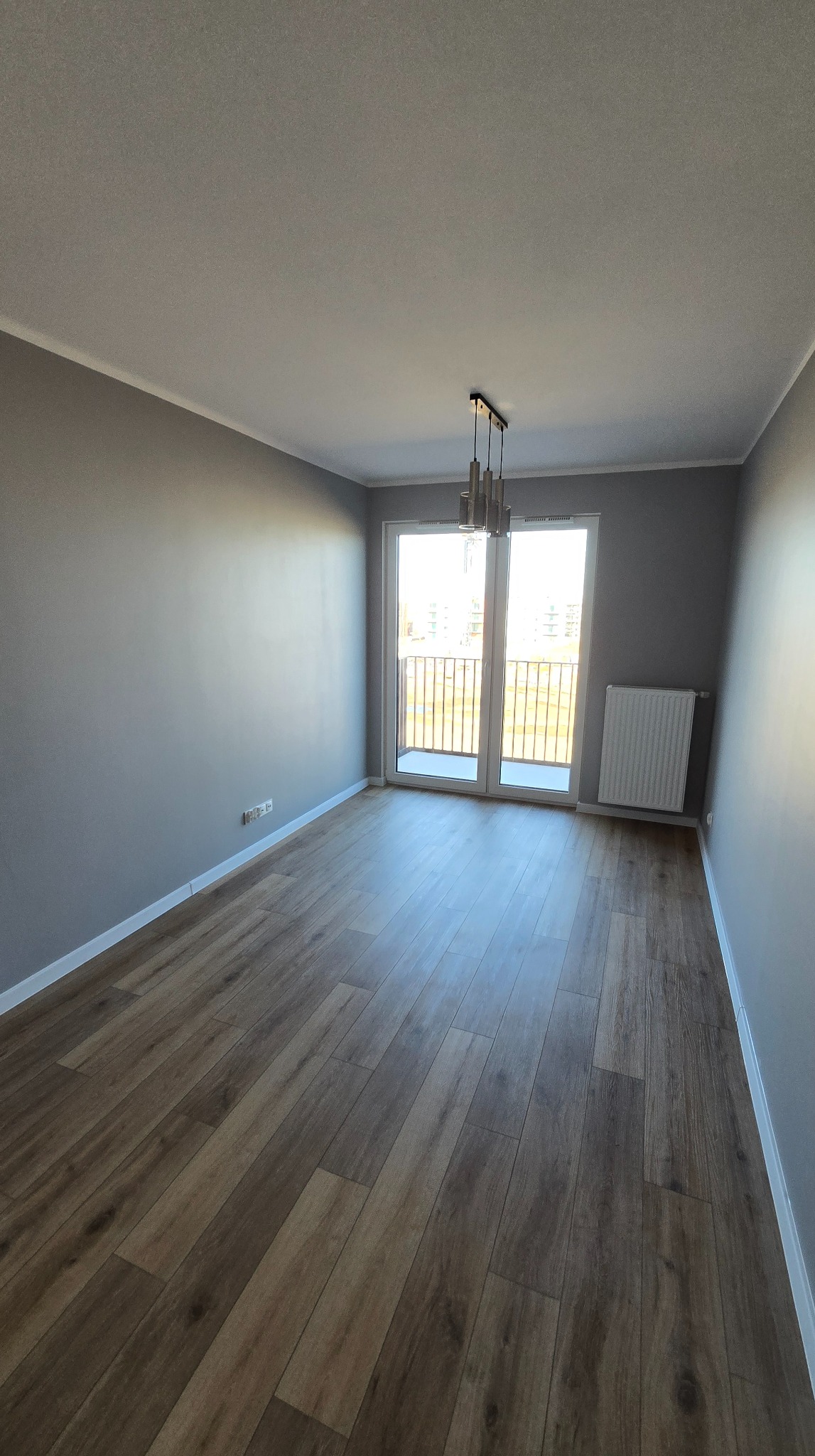 Jasne, przestronne wnętrze z drewnianą podłogą, szarymi ścianami i balkonem. Nowoczesna lampa wisząca dodaje elegancji. Widok na zewnątrz przez przeszklone drzwi balkonowe.
