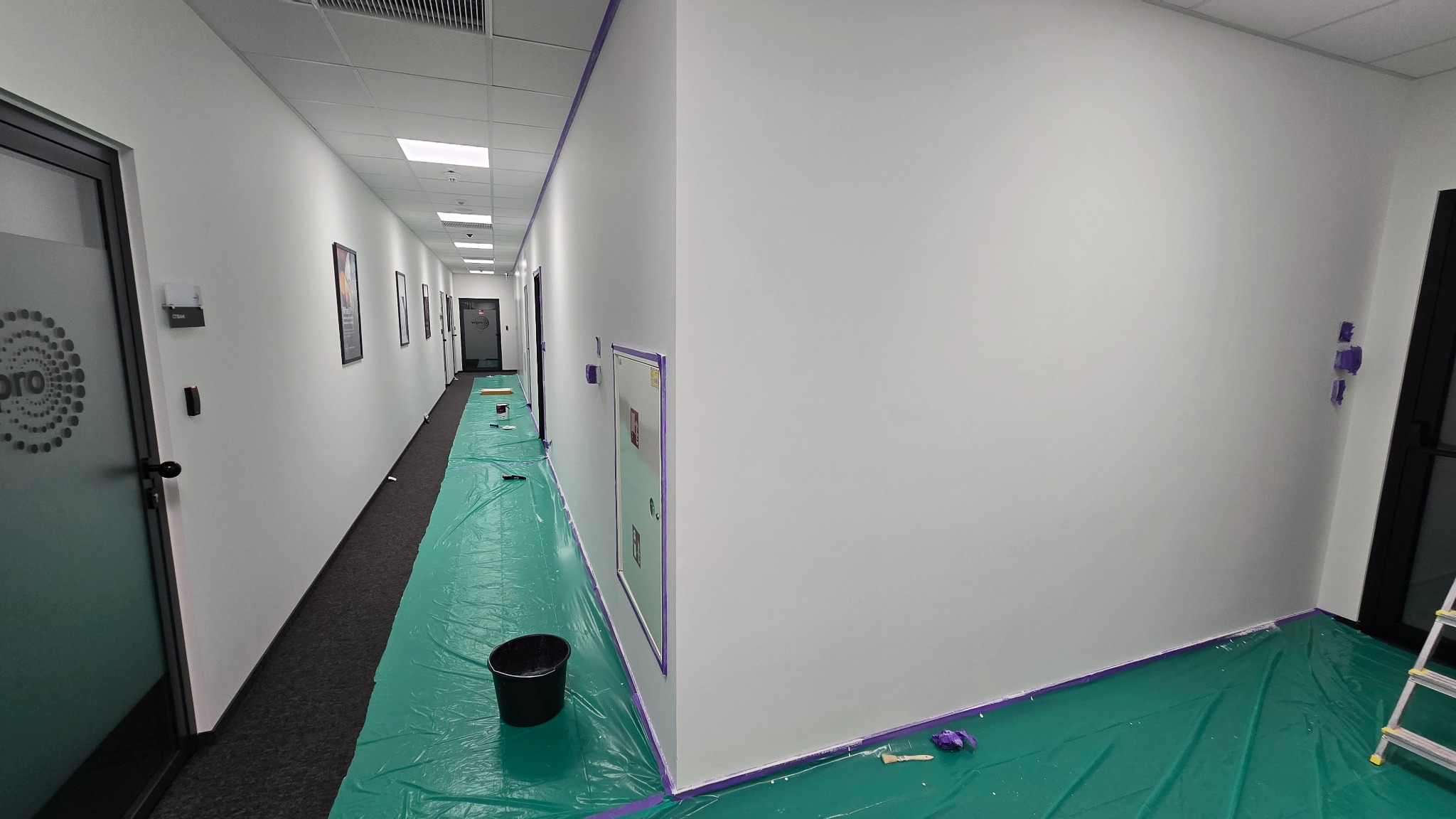Korytarz biurowy w trakcie malowania: zabezpieczona podłoga folią, świeżo pomalowane ściany na biało, taśma malarska wzdłuż krawędzi. Widoczny kubeł z farbą i drabina.