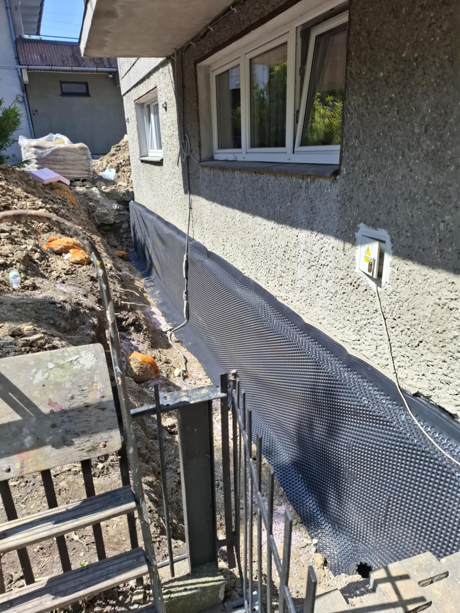 Częściowo odkryty fundament domu z ułożoną czarną folią izolacyjną. Widoczny wykop, schody i fragment ściany budynku z oknem.