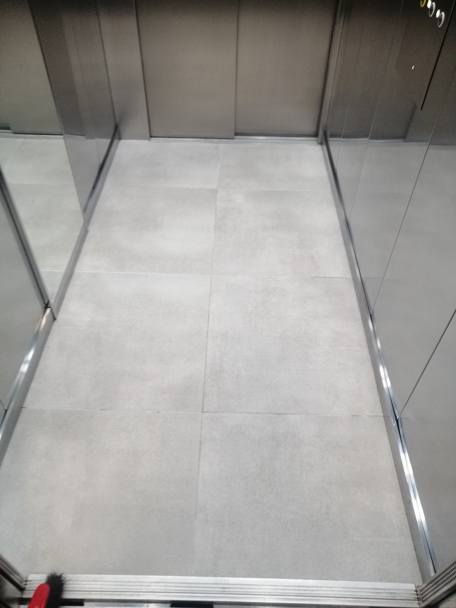 Wnętrze windy z szarymi płytkami podłogowymi, widok z góry na czystą, odświeżoną podłogę po doczyszczeniu. Metalowe ściany i listwy przypodłogowe.