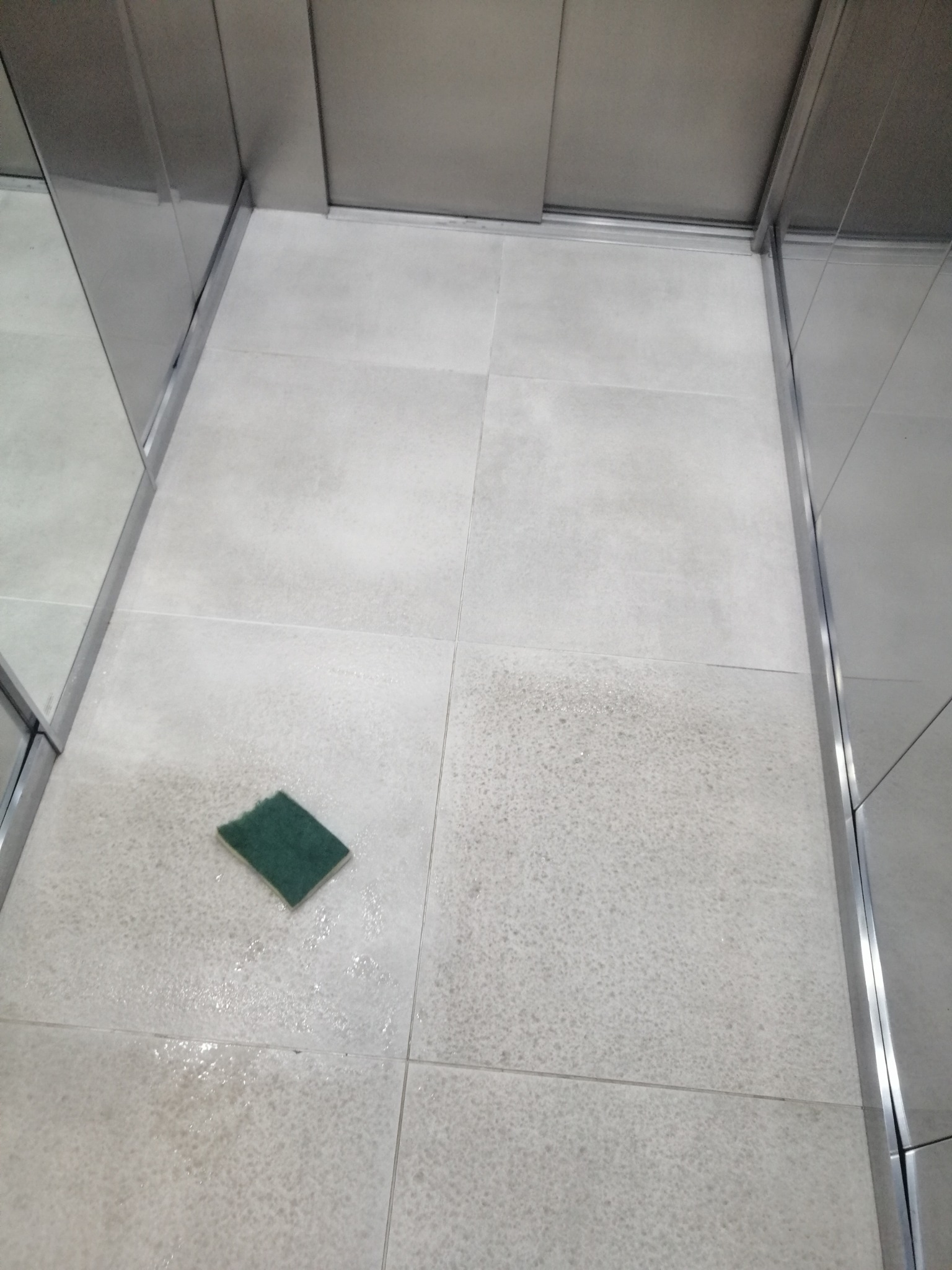 Wnętrze windy: mokra, jasna podłoga z płytek po umyciu, zielona gąbka leży na podłodze. Widoczne metalowe elementy konstrukcyjne windy i ślady wody.