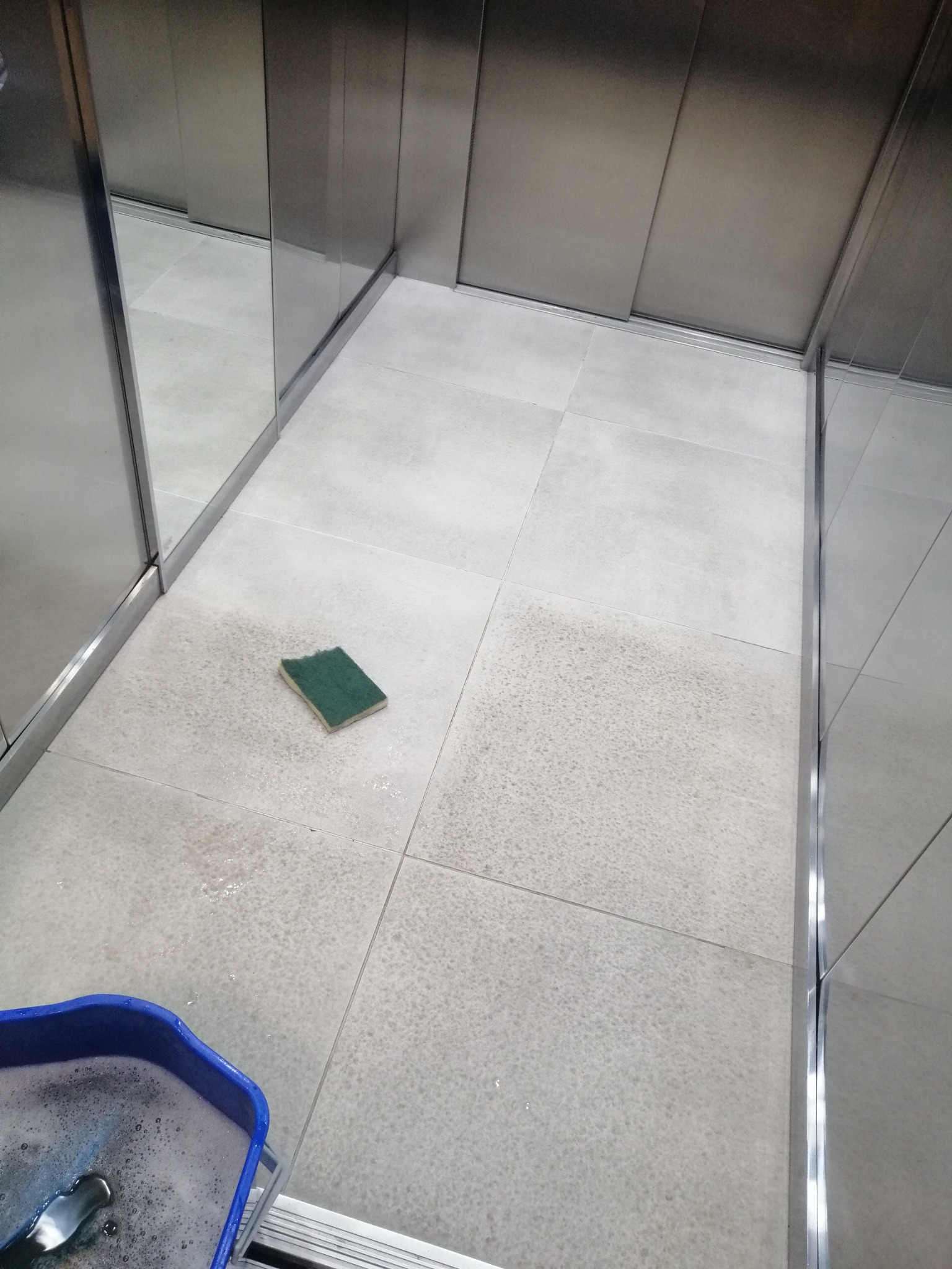 Czyszczenie podłogi w windzie: gąbka na mokrych, szarych płytkach, wiadro z pianą w rogu, metalowe ściany windy. Widok z góry, blisko podłogi.