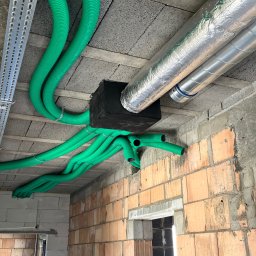 ALEKLIMA Alex Mozgała - Montaż instalacji wentylacyjnej z zielonymi rurami i srebrnymi przewodami w surowym wnętrzu budynku z ceglanymi ścianami. Widok z dołu na sufit.