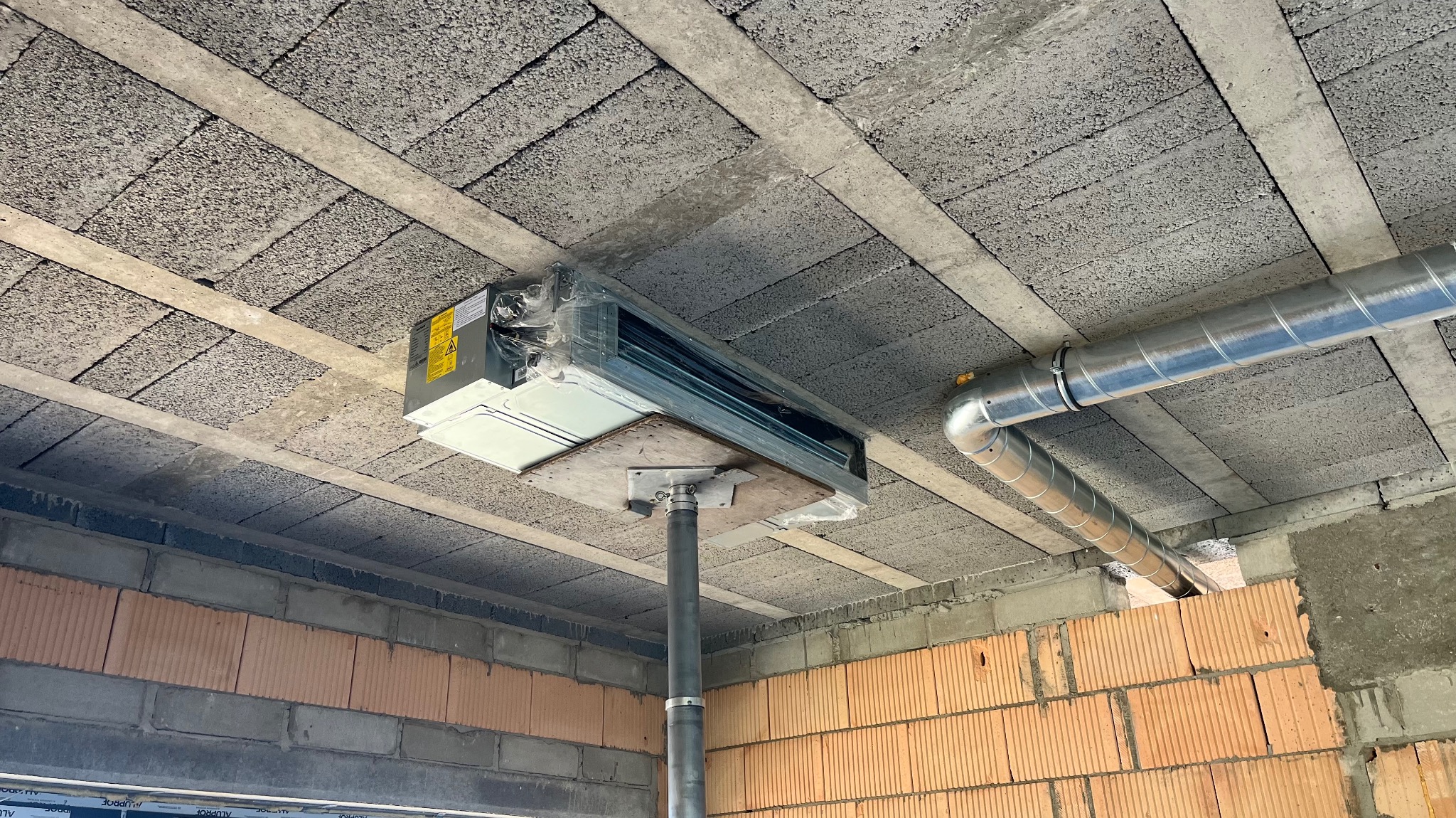 Montaż klimatyzacji kasetonowej pod stropem z pustaków, widoczne rury wentylacyjne i fragment ściany z cegieł. Klimatyzator jeszcze w folii ochronnej.