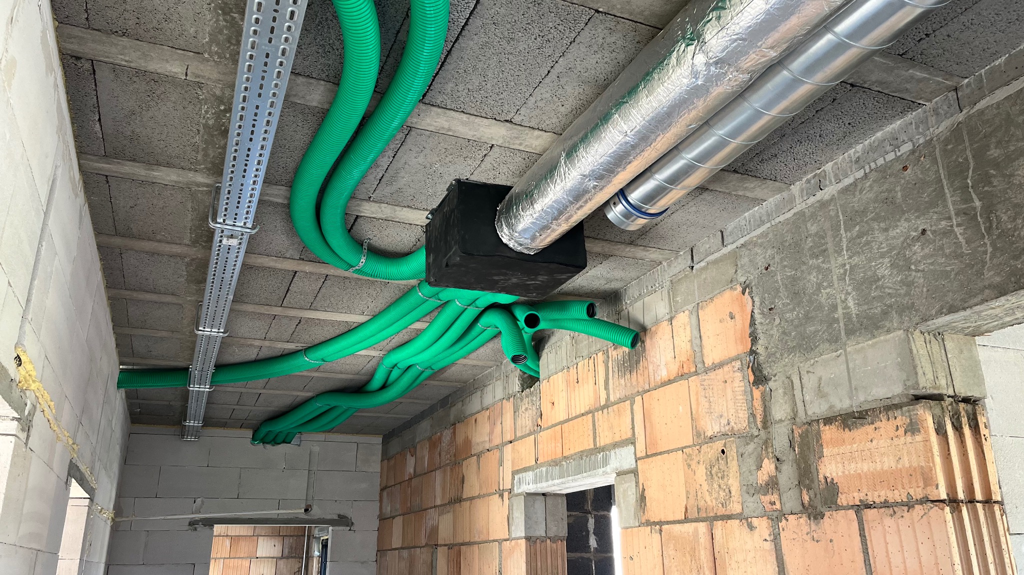 Montaż instalacji wentylacyjnej z zielonymi rurami i srebrnymi przewodami w surowym wnętrzu budynku z ceglanymi ścianami. Widok z dołu na sufit.