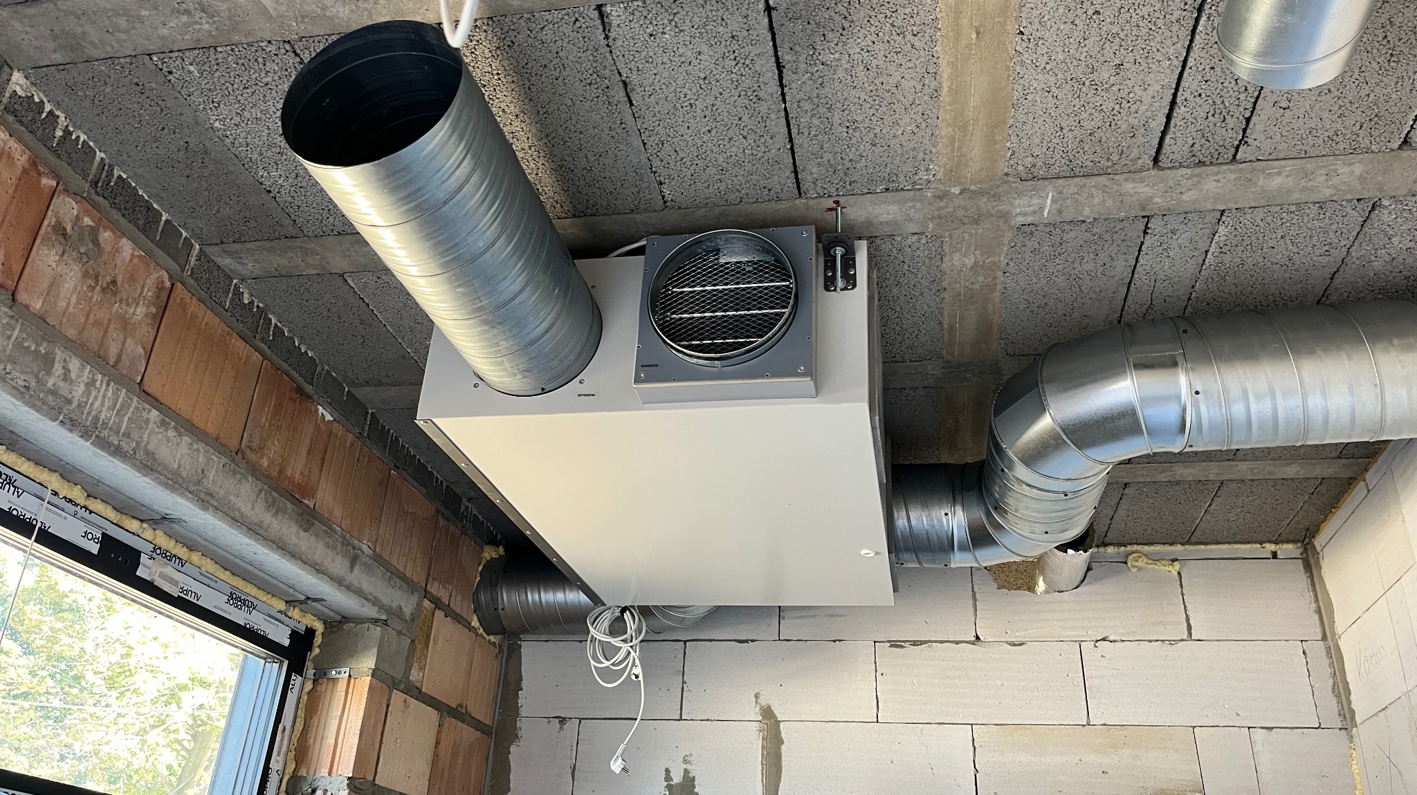 Centrala rekuperacyjna z widocznymi kanałami wentylacyjnymi, zamontowana pod betonowym stropem w surowym wnętrzu budynku. Widoczne okno w stanie deweloperskim.